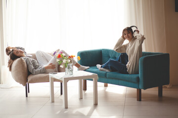 Two  teenagers girls  is relaxing at home in the living room in headphones, sitting in an armchair and sofa,  listening music in headphones
