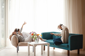 Two  teenagers girls  is relaxing at home in the living room in headphones, sitting in an armchair and sofa,  listening music in headphones