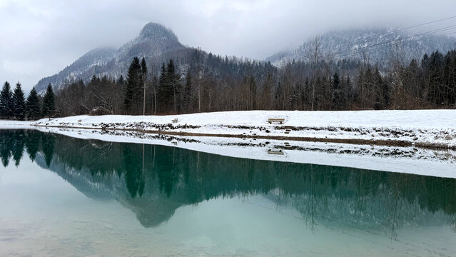 A tranquil alpine lake reflects snow-covered mountains and evergreen trees on a winter day. The turquoise water, misty peaks, and wooden benches create a serene and picturesque nature landscape.