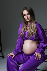 Obraz premium Portrait of attractive pregnant woman in violet costume, trousers, studio photoshoot. Fashion photo of pregnancy. Young pretty girl with long hair touching her belly. Isolated, grey background