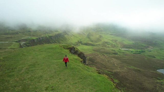 Aerial Hiking Girl Walks Green Lush Scottish Highlands Quiraing Foggy Morning. Female Model Orange Jacket Walks to Cliff Drone Past Her to Moody Landscape