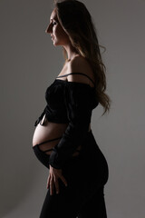 Fototapeta premium Portrait of attractive pregnant woman in black costume, trousers, top, studio photoshoot. Fashion photo of pregnancy. Young pretty girl with long hair touching her belly. Isolated, grey background