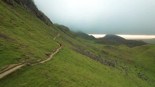 Epic Drone Follows and Passes Female Photographer Hiking Remote Trail in Scottish Highlands. Orange Coat Girl Walks the Quiraing Cloudy Morning Aerial