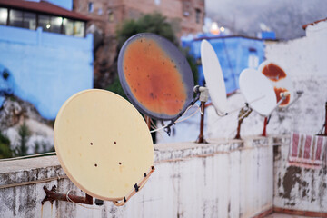 A row of weathered satellite dishes mounted on a rooftop, with a backdrop of blue-painted buildings.