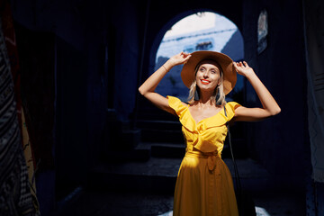 A woman in a bright yellow dress and wide-brimmed hat stands in a dark, arched alleyway, smiling...