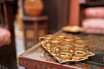 Fototapeta premium A set of ornate brass tea cups on a decorative wooden table, with a blurred background of traditional furniture and decor, creating a warm and inviting atmosphere.