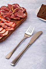 Sliced cured meats arranged on rustic wooden board with fork and knife on gray surface. Piece of dark rye bread on plate adds contrast. Concept of traditional charcuterie and gourmet dining