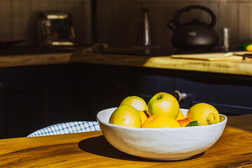 A plate of fresh seasonal fruit stands on a wooden table in a rustic cozy stylish kitchen. Healthy food, nutrition, vitamins for a healthy lifestyle. Apples, oranges, lemons, citrus on a platter.