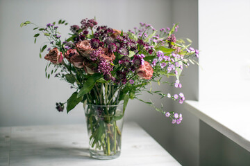 Wedding bouquet of ash-pink roses, Great Masterwort, Astrantia, Milkweed Plant, and Hooked Hakea in purple and pink tones near a window on a beige table with a white background. Space for text.