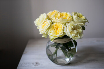 Bouquet of white-lemon garden tea roses in a glass vase on a beige table against a white wall background with space for text.