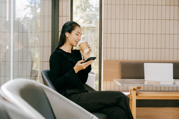 Serene Coffee Break: An Asian woman, lost in thought while sipping coffee and engaging with her phone. Captured inside a modern setting with large windows.