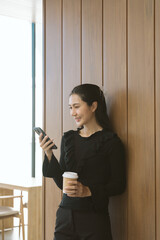 Digital Moment: An elegant woman pauses in a modern space, engrossed in her phone, a warm drink in...