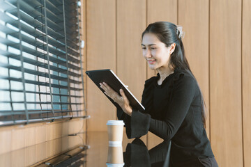 Digital Engagement: A businesswoman, engrossed in her tablet, stands near a window, with a takeaway...