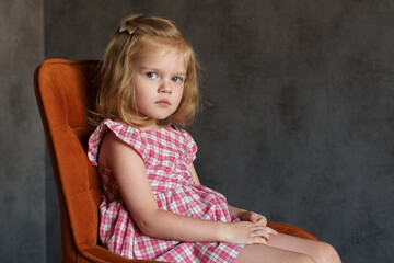 Fototapeta premium Little girl sitting on a orange chair in checkered dress, white bow in the hair looking seriously 