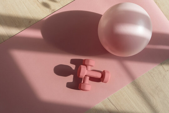 Pink pilates ball and dumbbells on a pink sport mat, prepared for a home workout, soft sunlight from the window conveys the atmosphere of the morning