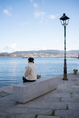 Tourist sitting on concrete bench, overlooking tranquil waters and distant hills of combarro, absorbing serene coastal landscape of galician seaside promenade