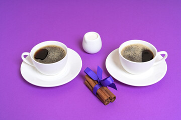 Two Coffee Cups with Cinnamon Sticks and Milk Jug on a Purple Background