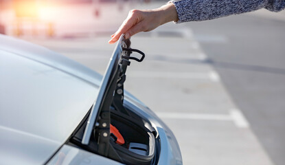 A person wearing a gray sweater opening the charging port cover of a white electric car on a...