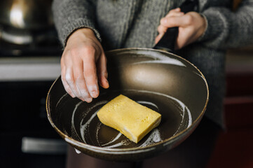 Diligent adult woman housewife washes, cleans with yellow sponge and detergent with foam dirty teflon round frying pan, dishes in kitchen. Close-up photo, hygiene and cleaning concept.