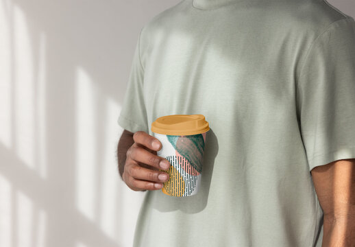 Anonymous man stands indoors holding a disposable takeaway paper cup with a lid,  PSD template mockup ready for your Design