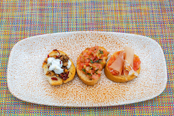 Three assorted bruschetta appetizers with toppings on ceramic plate over colorful woven table matю Mexico.
