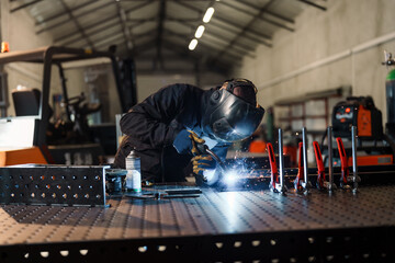 Welder wearing protective gear is using a welding torch to join metal pieces on a welding table in a factory, creating sparks and bright light, demonstrating industrial manufacturing processes