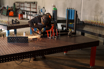 Metalworker wearing protective gear uses an angle grinder to smooth a steel frame clamped to a...
