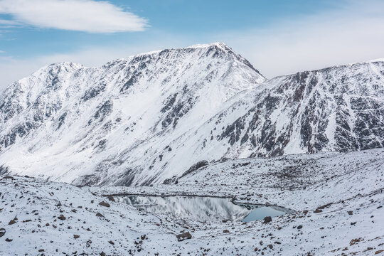 Cold snow-white landscape of snowy valley with alpine lake against snow-covered rocky mountain range under clouds in blue sky. Glacial lake, pure white snow and stones in high mountains in sunny day.
