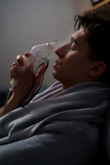 Sick man wrapped in a blanket is doing inhalation by nebulizer for the cough treatment. Patient is...