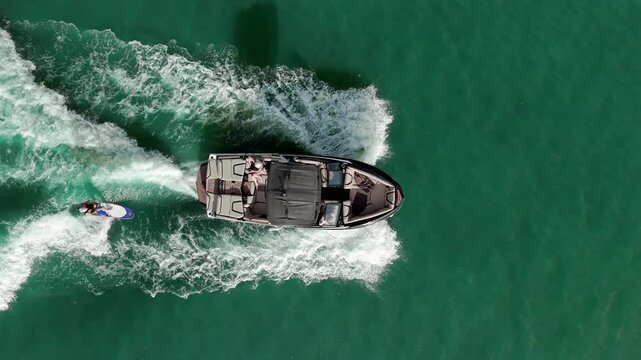 Aerial view of a speedboat moving across a vibrant water. A person is being towed behind the boat, wakeboarding or water skiing, Carving through the water with a dynamic posture.
