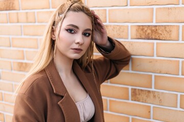 Young blonde woman with stylish braids, wearing a chic brown coat, posing confidently against a textured brick wall, showcasing her elegant fashion sense and natural beauty