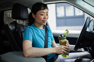 Woman in sportswear sitting in car drinking water after workout