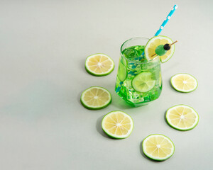 Mint cocktail garnished with lemon slice and green cherry surrounded by lemon slices on white table