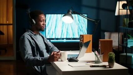 Young casual guy listening to a podcast while he works on freelance job at home, enjoys multitasking with headphones. Man solving tasks and paying attention to webinar discussion. Camera B.
