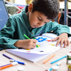 Smart Indian little boy perform thumb painting with different colourful water colour kit during the summer vacations, Cute Indian Kid doing colourful thumb painting drawing on wooden table