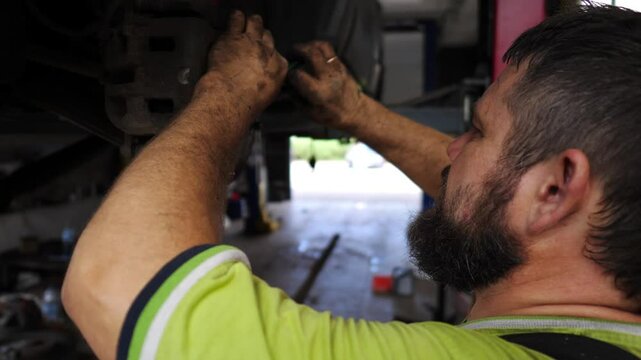 Male auto mechanic in uniform works underneath a lifting vehicle at workshop. Adult professional repairman fixing car at service or garage. Concept of regular automobile maintenance. Slow motion