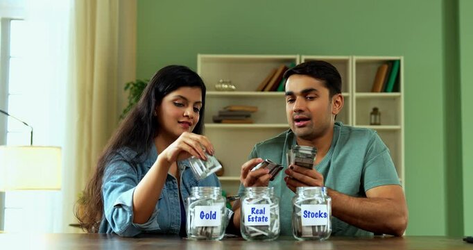 Indian Asian young couple in twenties at home desk, placing Indian currency notes into three glass jars labeled Gold, Real Estate, Stocks, symbolizing financial planning, investments, Future security