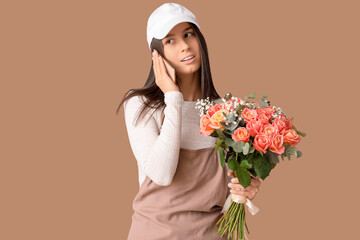 Young delivery woman with bouquet of beautiful flowers talking by mobile phone on brown background