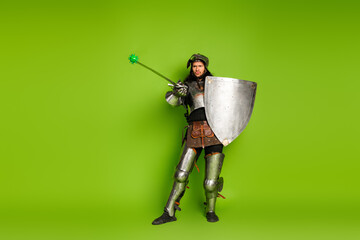 Medieval knight in full armor holding shield and wand against vibrant green background,...