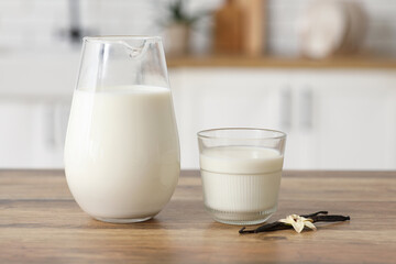 Glass and jug of fresh vanilla milk with sticks and flower on wooden table in kitchen