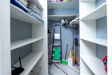 A small, clean, and organized closet with a ladder and a broom leaning against the wall. The closet is filled with clothes and other items, but it is well-organized and tidy