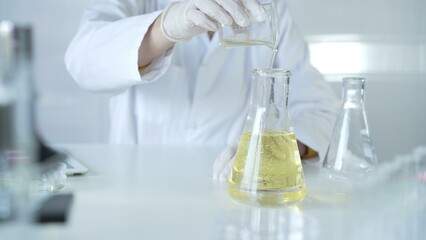 A scientist, wearing a lab coat and white protective gloves, is pouring a yellow oily liquid from one beaker to another in laboratory, close up. Medicine and science concept