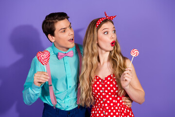 Young couple in retro-style clothing holding lollipops on a vibrant purple background, expressing joy and charm