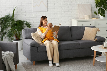 Young happy mother with little baby in sling reading book at home