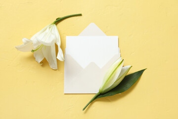 Composition with white lily flowers and blank card in envelope on yellow background. Top view