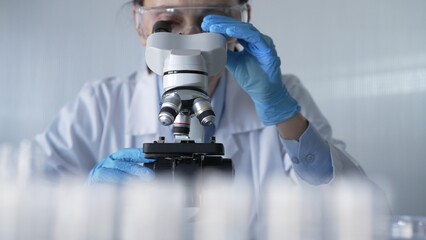 Woman scientist wearing lab coat, white protective gloves and glasses, is using microscope in...
