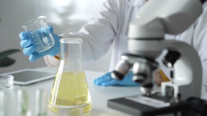 Scientist, wearing blue protective gloves, is holding a beaker with yellow liquid while sitting at the table in laboratory besides microscope, close up. Medicine and science concepts