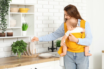 Young mother with little baby in sling taking plate in kitchen