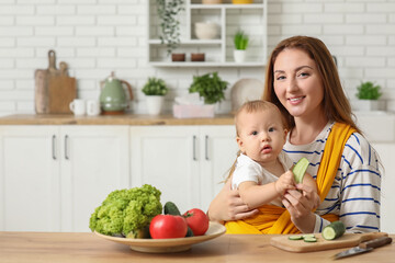 Young mother with little baby in sling and cucumber in kitchen