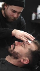 Obraz premium Professional barber carefully shaving client's beard with straight razor, applying gel and using hot towel for precise grooming in contemporary barbershop setting, vertical view